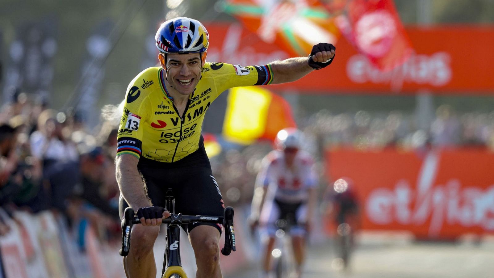 Ciclista con maillot amarillo y número 13 celebra victoria, puño en alto.  Logos de patrocinadores visibles en la equipación. Público y publicidad de fondo.