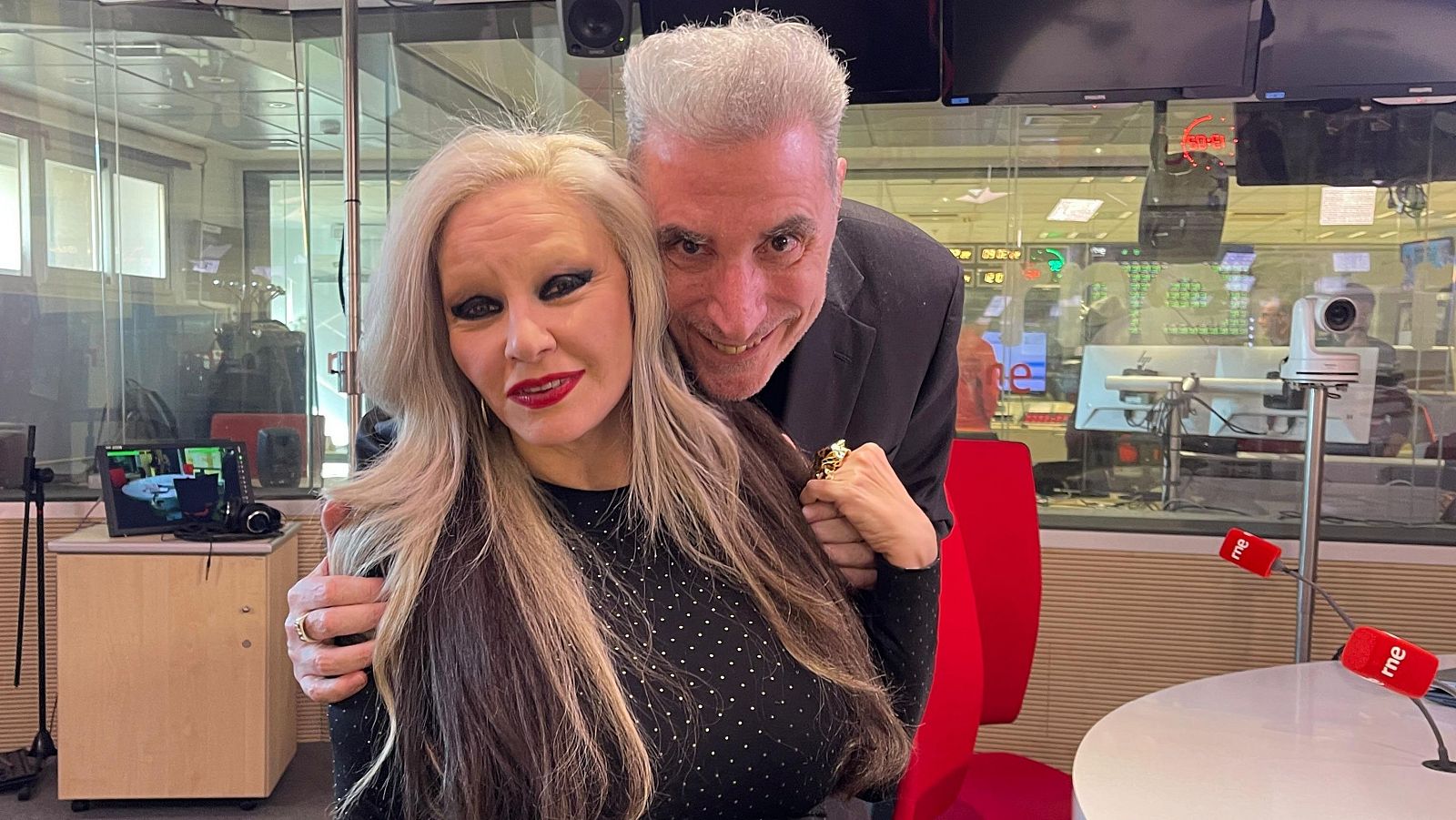 Alaska y Loquillo, sonrientes y abrazados, en un estudio de radio con micrófonos y una mesa redonda.  Se ve una sala de control tras una pared de cristal.