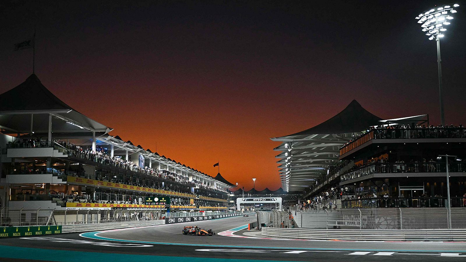 Monoplaza de F1 en curva nocturna en el circuito Yas Marina.  Gradas llenas, iluminación artificial y estructuras arquitectónicas visibles.