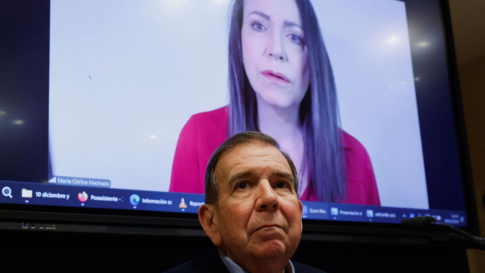 Videoconferencia: Un hombre observa con atención a una mujer en una pantalla, mostrando una fecha del 10 de diciembre. La mujer viste una blusa roja y tiene expresión seria.