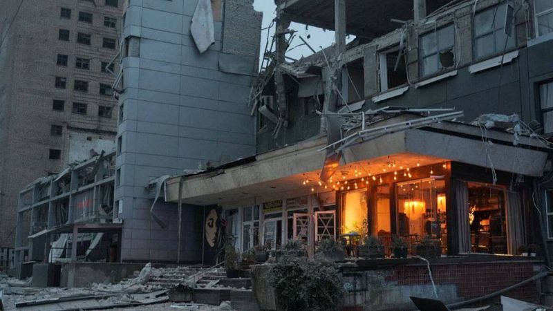 Edificio multiplanta severamente dañado, con escombros, ventanas rotas y un local iluminado en la planta baja. Grafiti visible en una pared.