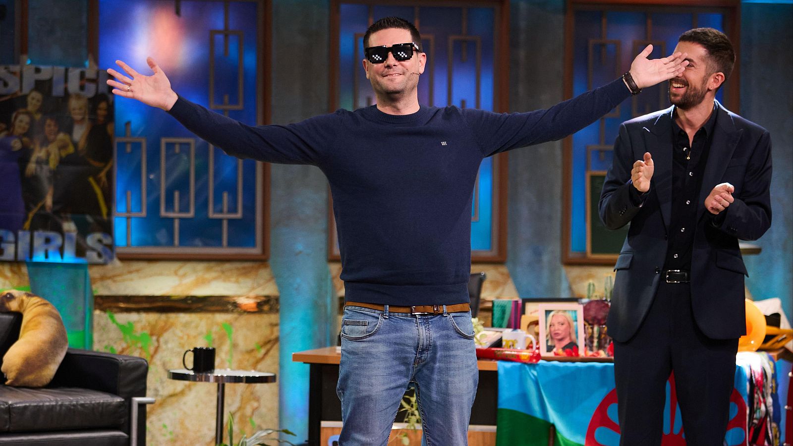 Dos hombres, uno con jersey azul y gafas, el otro con traje, interactúan en un plató de televisión. Uno celebra o saluda, el otro sonríe con complicidad.