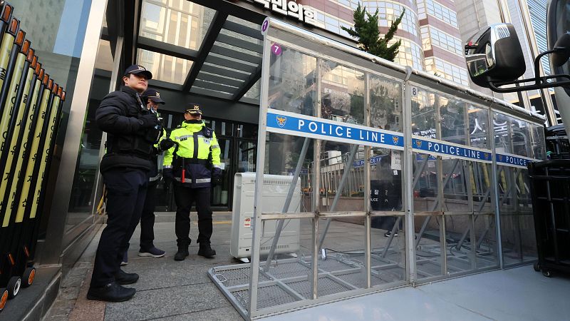 Agentes de policía custodian la entrada a un edificio, con una barrera transparente que indica "POLICE LINE" y barreras metálicas plegables adicionales.