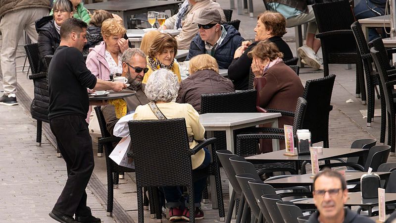 Terraza de bar con clientes sentados en mesas oscuras, un camarero atiende a un grupo. Ambiente de conversación y consumo.