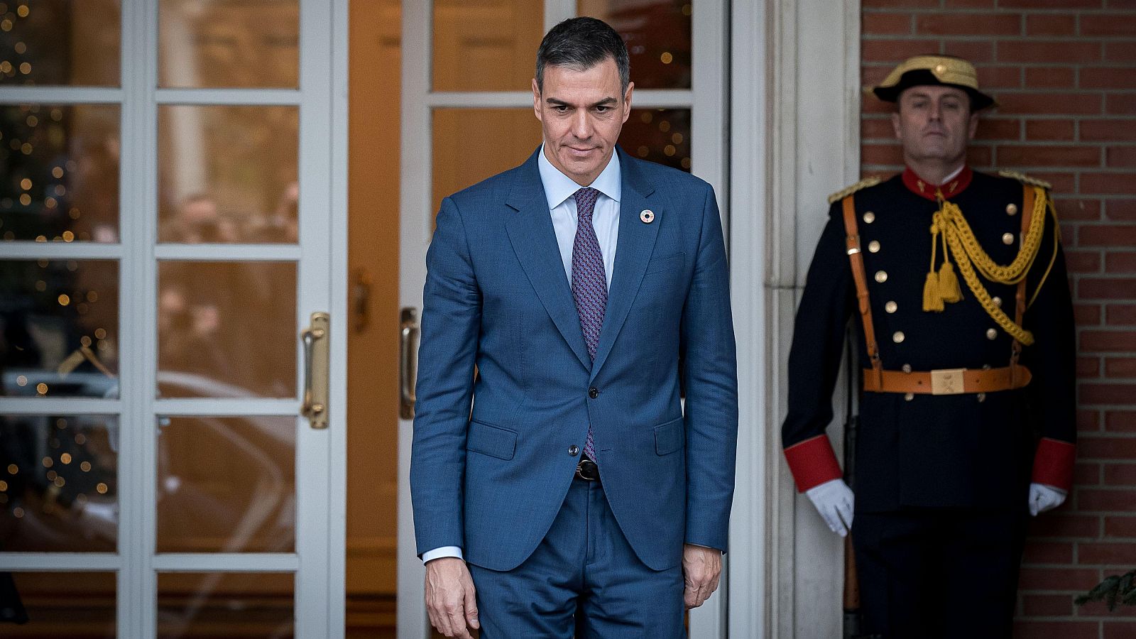 El presidente del gobierno español, Pedro Sánchez, en la entrada de un edificio, acompañado por un guardia de seguridad con uniforme. Sánchez viste traje azul marino y corbata morada.