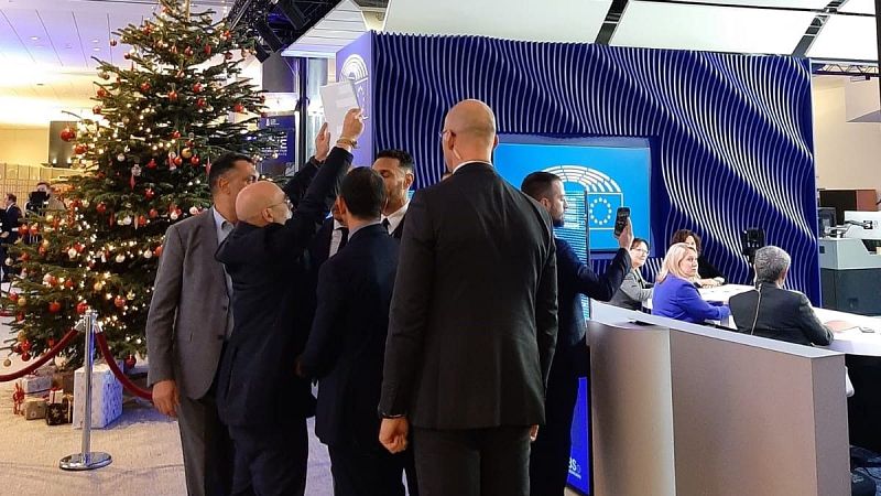 Evento en el Parlamento Europeo: personas con móviles grabando, árbol de Navidad decorado visible y panel con logo de la UE al fondo.
