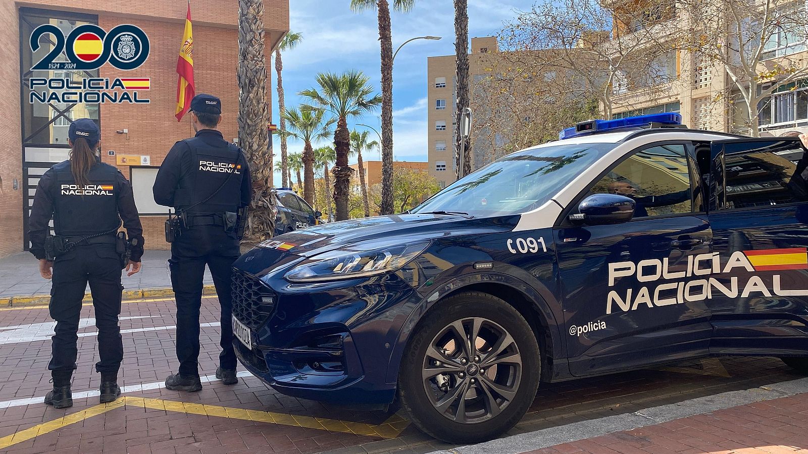 Agentes de policía en uniforme frente a una comisaría con coche patrulla y banderas españolas.  Logo conmemorativo del bicentenario policial visible en el edificio.