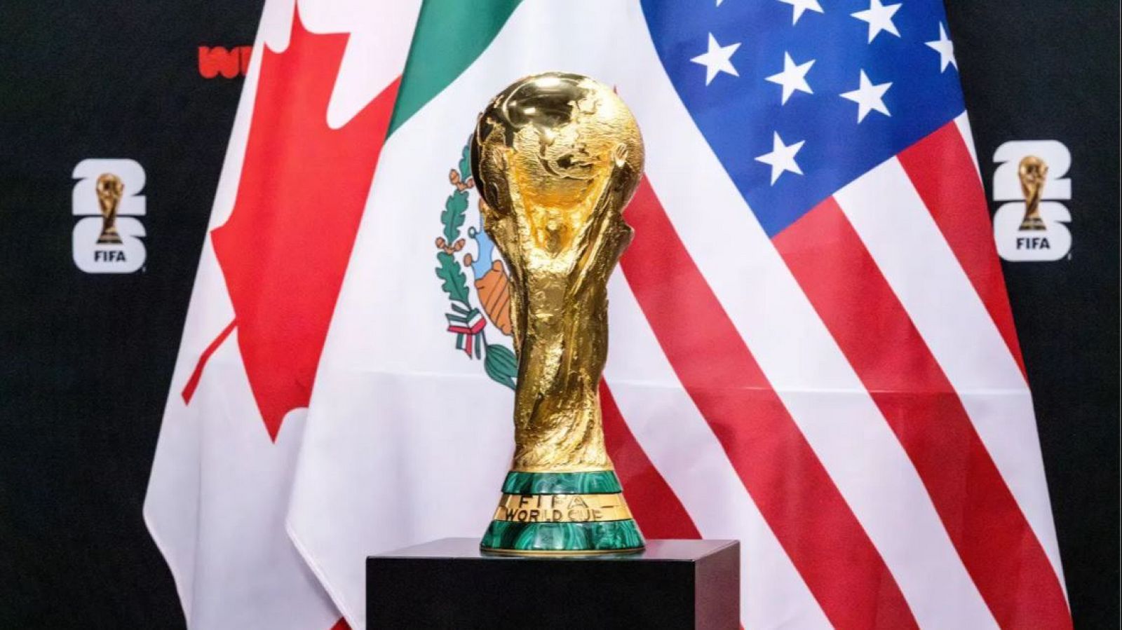 Trofeo de la Copa Mundial sobre pedestal con banderas de Canadá, México y EEUU al fondo.  Imagen relacionada con el sorteo del Mundial 2026.