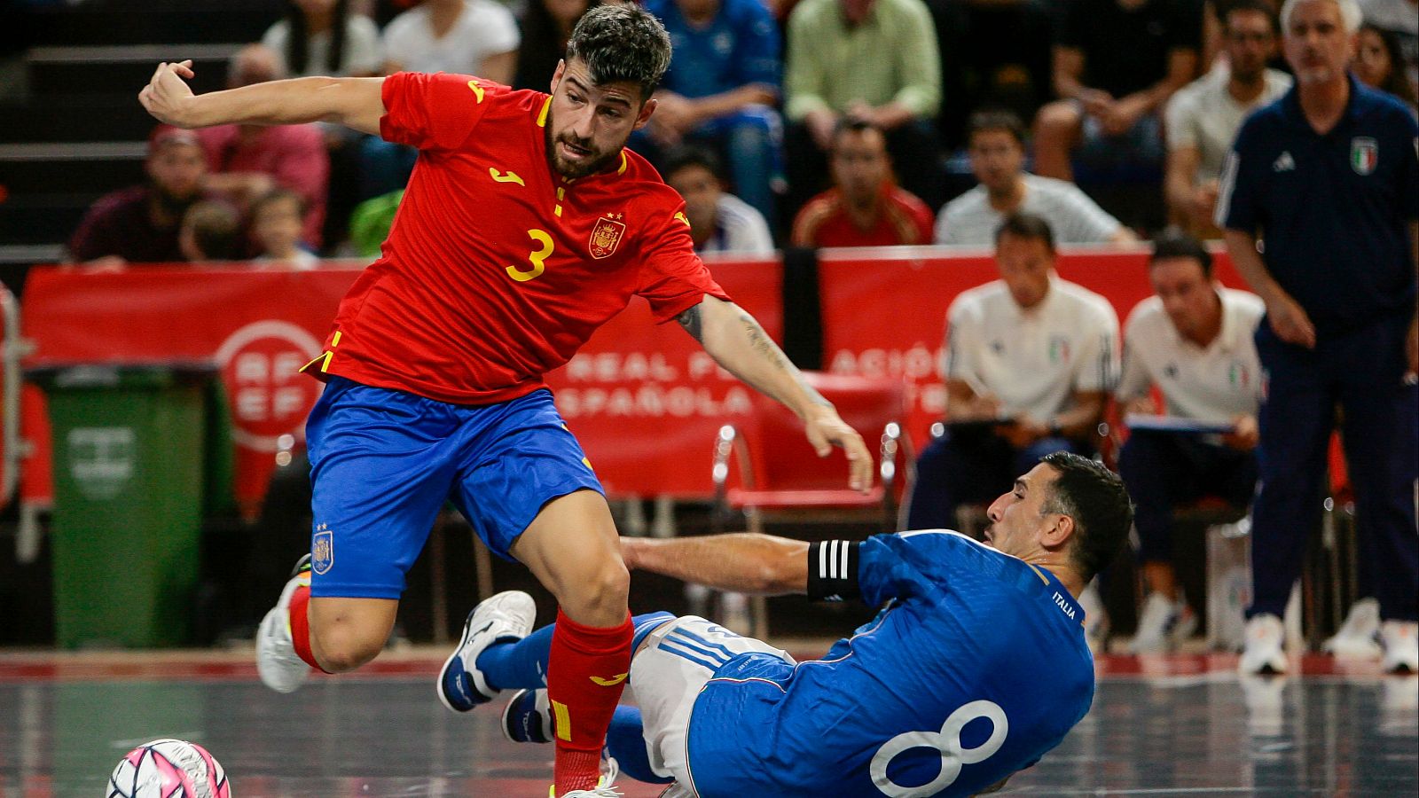 Partido de fútbol sala: jugador español (camiseta roja, #3) regatea, dejando en el suelo a un jugador italiano (#8). Balón rosa y blanco visible.