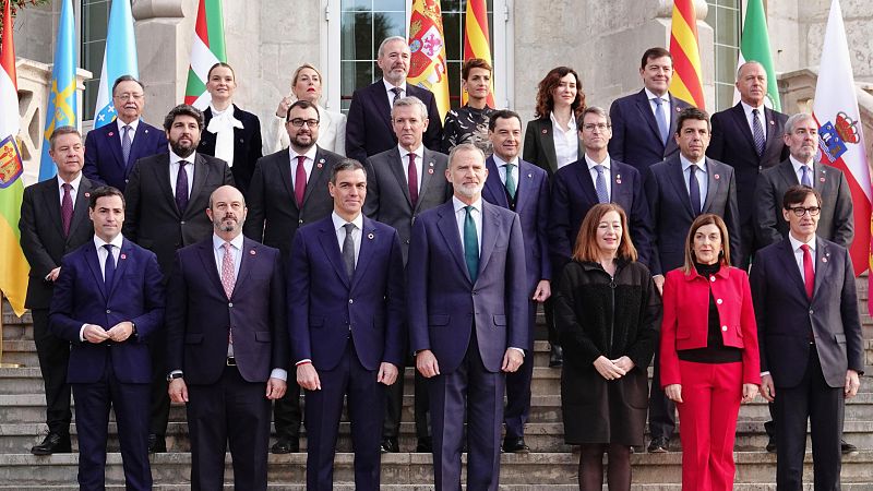 Grupo de personas en trajes formales, incluyendo al Rey Felipe VI y Pedro Sánchez, en una escalera.  Banderas de fondo. Evento formal.