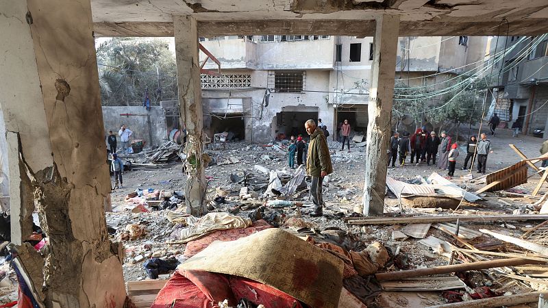 Destrucción en un campo de refugiados: escombros, personas inspeccionando daños y edificios con paredes derruidas.  Se observa una posible mancha de sangre.