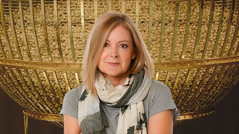 Imagen promocional de Pepa Fernández presentando el especial de la Lotería de Navidad 2024 en RNE. Lleva camiseta gris y bufanda estampada; se ve el bombo de la lotería.