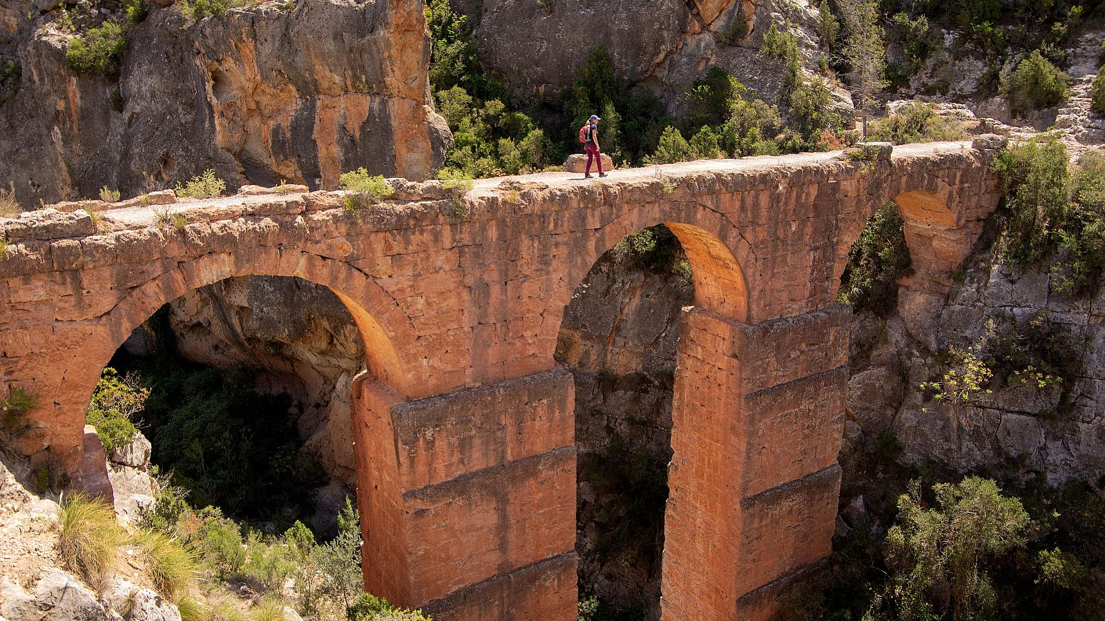 Impresionantes restos de un acueducto de piedra rojiza con múltiples arcos, sobre terreno rocoso. Una persona con mochila camina por la parte superior.