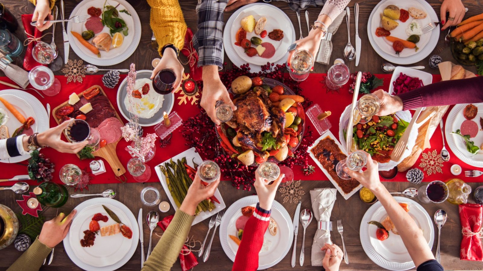 Mesa navideña con mantel rojo, pavo asado y otros manjares, rodeada de comensales vestidos para la ocasión.