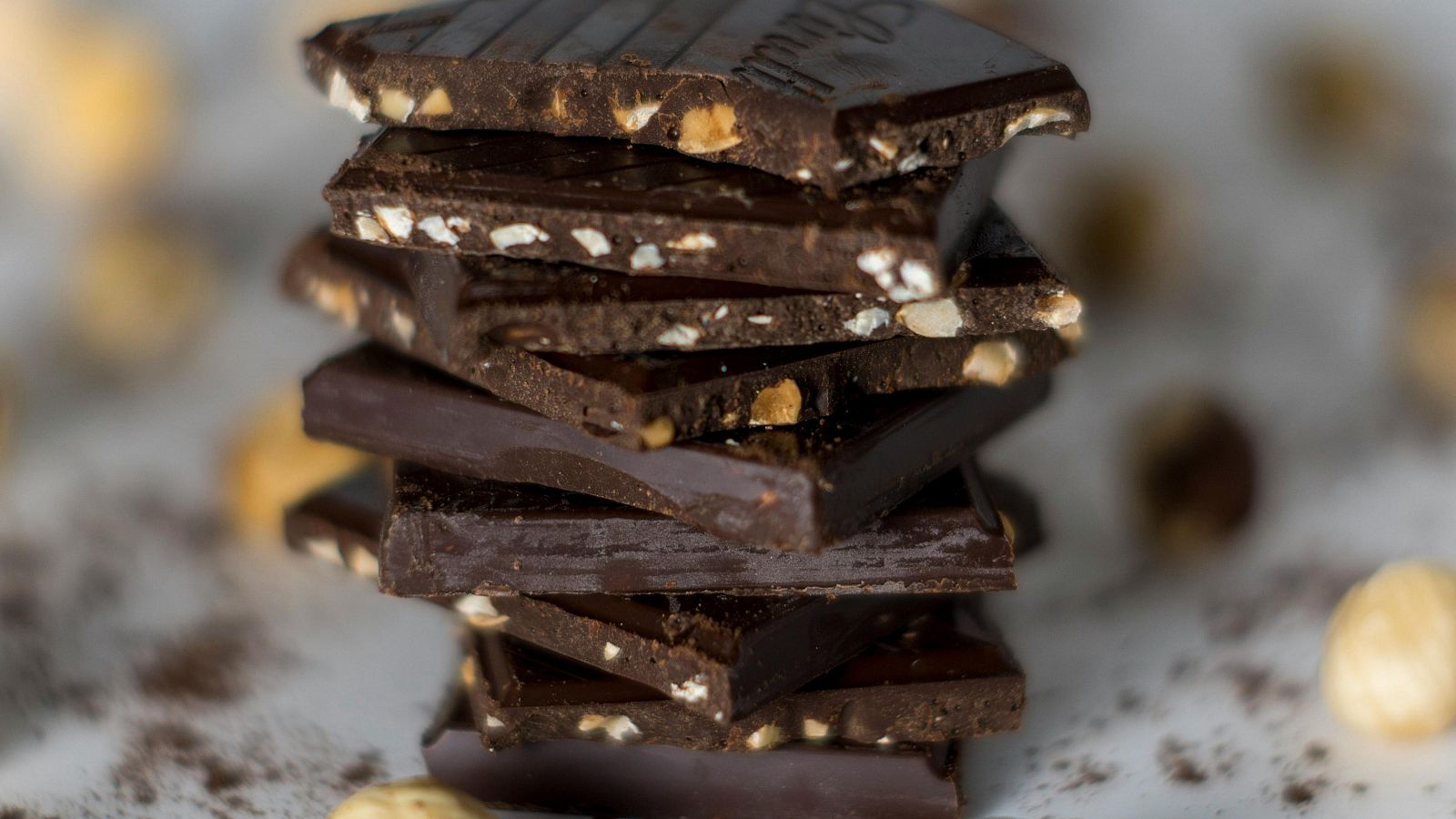 Trozos de chocolate negro con avellanas, algunos rotos, sobre un fondo desenfocado con avellanas y cacao en polvo.