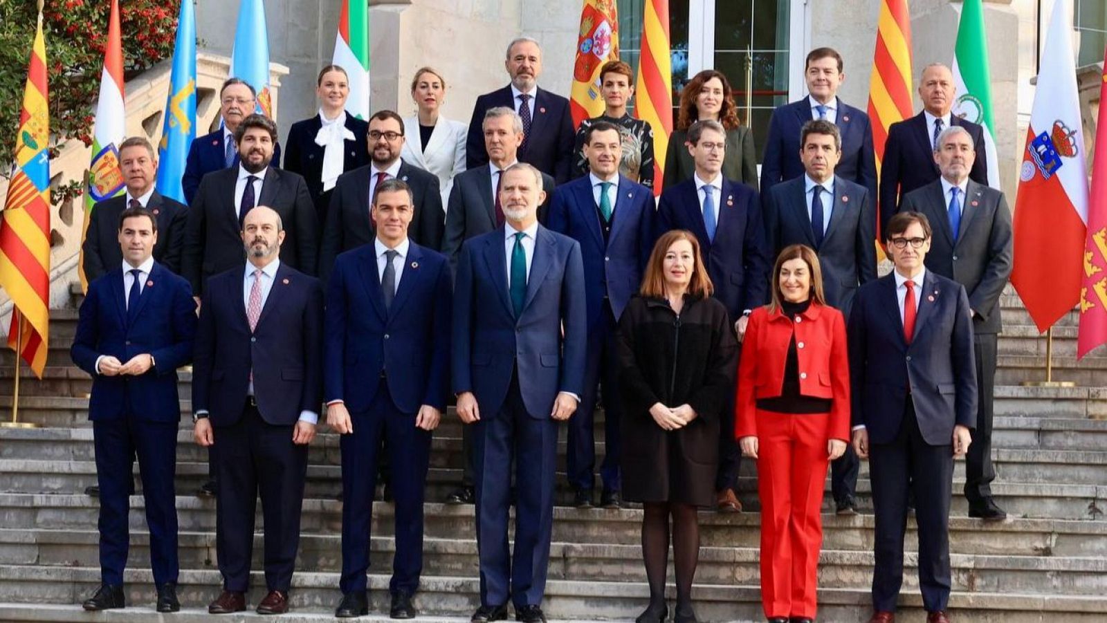 Grupo de personas, mayoritariamente hombres en traje y mujeres con trajes de chaqueta o vestidos, posan en una escalinata. El Rey Felipe VI se encuentra en el centro.