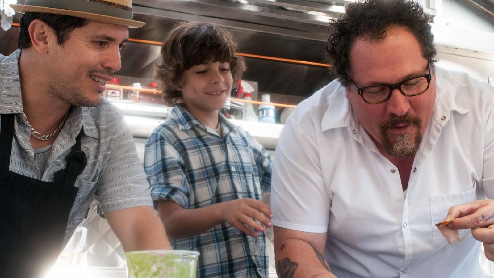 En la cocina de un food truck, Jon Favreau muestra comida a dos compañeros, uno con sombrero y delantal, el otro con camisa a cuadros.