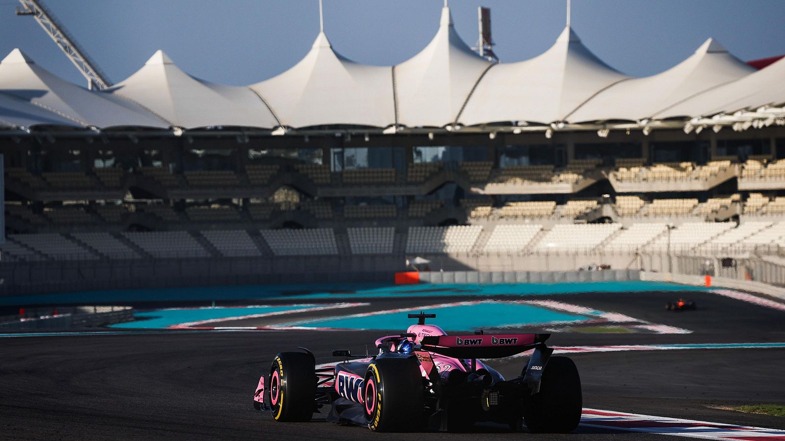 Monoplaza de F1 en carrera, colores rosa y negro, circuito con tramos turquesa, rosa y blanco; tribuna con techo de lona al fondo.