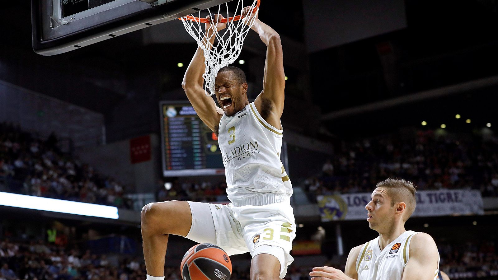 Mate impresionante de un jugador con camiseta blanca y el número 3 ('Palladium') durante un partido de baloncesto.  Otro jugador defiende cerca del aro.