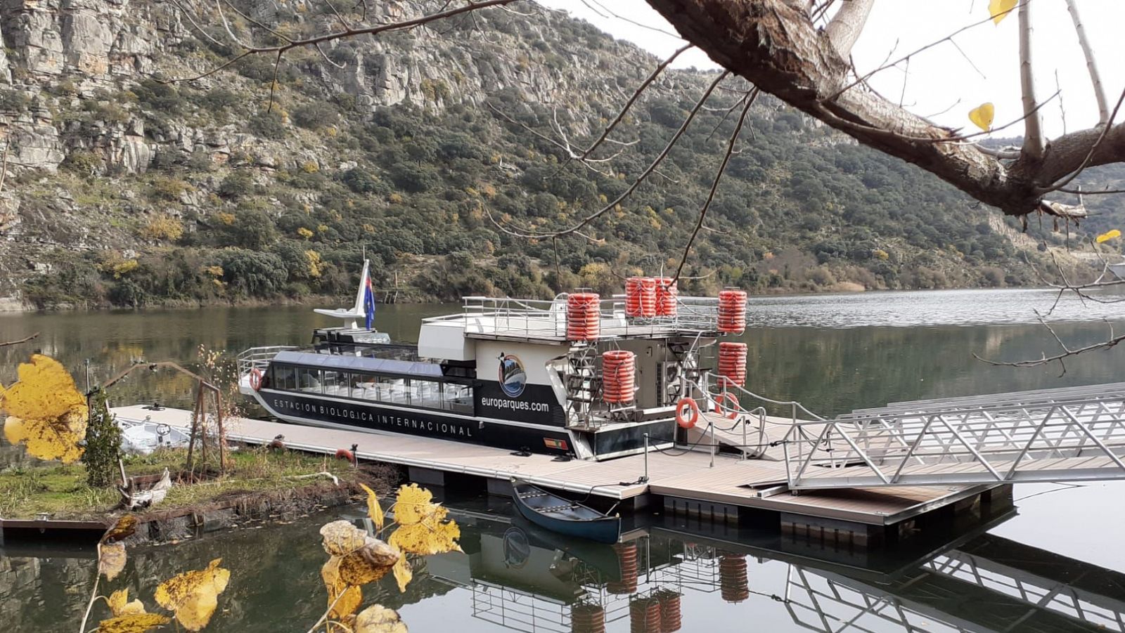 Crucero ecológico blanco y negro con dos cubiertas, equipamiento rojo y texto 'Estación Biológica Internacional'. Atracado en muelle con bote pequeño cercano, en entorno natural otoñal.