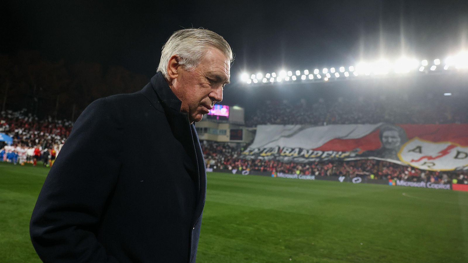Imagen de Carlo Ancelotti al borde del campo, con la cabeza baja.  Aficionados en la grada y la iluminación del estadio son visibles.  Contexto: tensión entre optimismo y expectativas.
