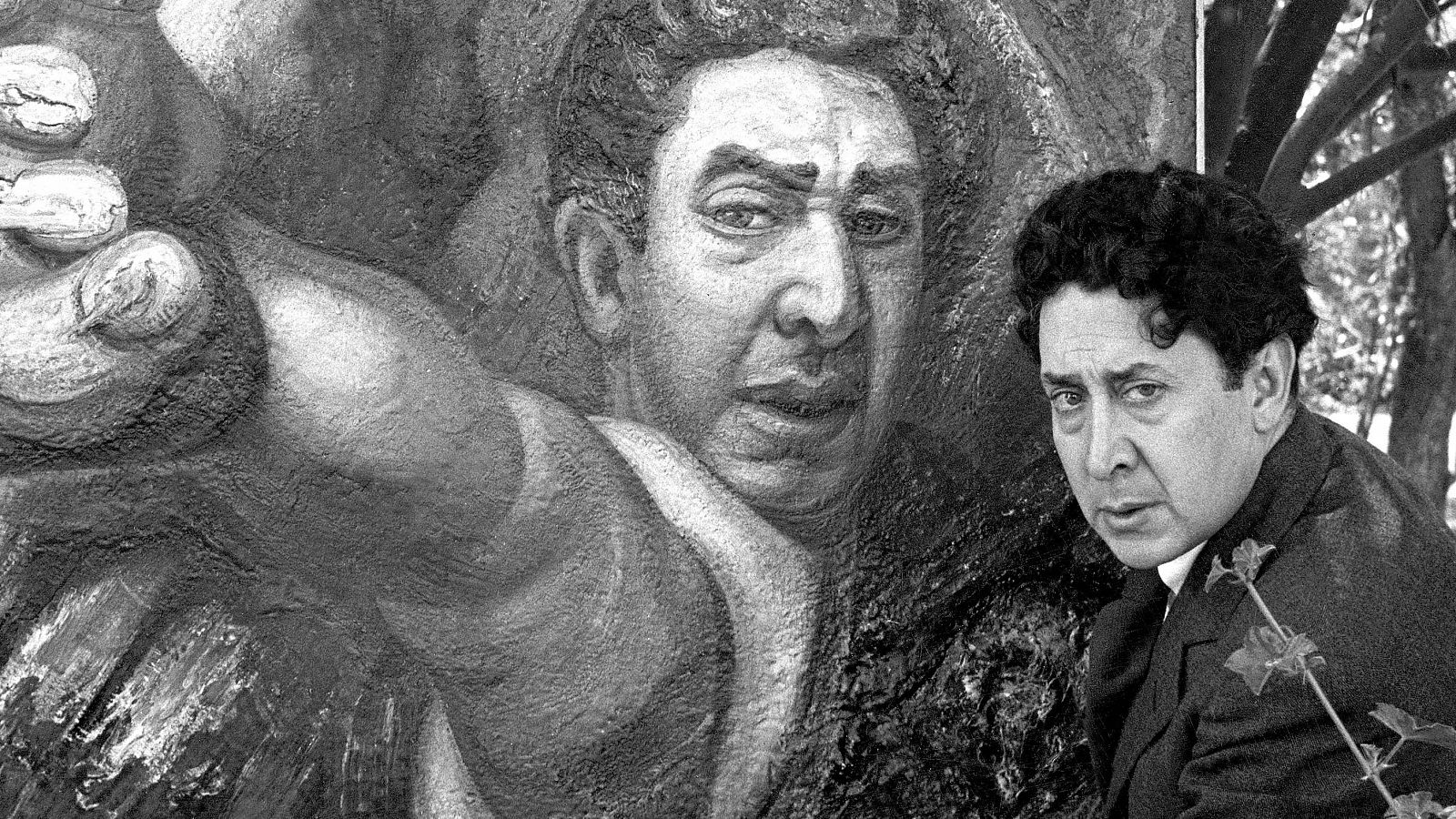 Autorretrato de David Alfaro Siqueiros en blanco y negro: medio cuerpo, traje oscuro, junto a un mural con figura masculina dinámica. Árbol visible al fondo.