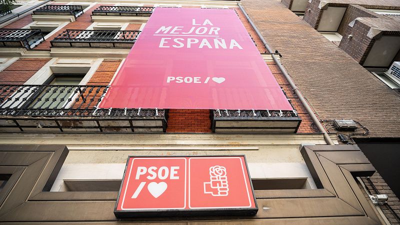 Fachada de edificio con cartel rosa gigante que dice "LA MEJOR ESPAÑA" en letras blancas y logo del PSOE repetido en la pancarta y cartel luminoso.