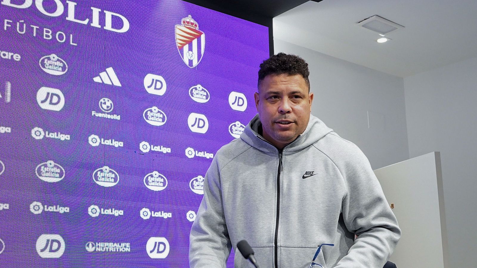 Rueda de prensa de Ronaldo Nazário con sudadera gris y logos de patrocinadores. Micrófonos presentes, indicando declaraciones a medios.