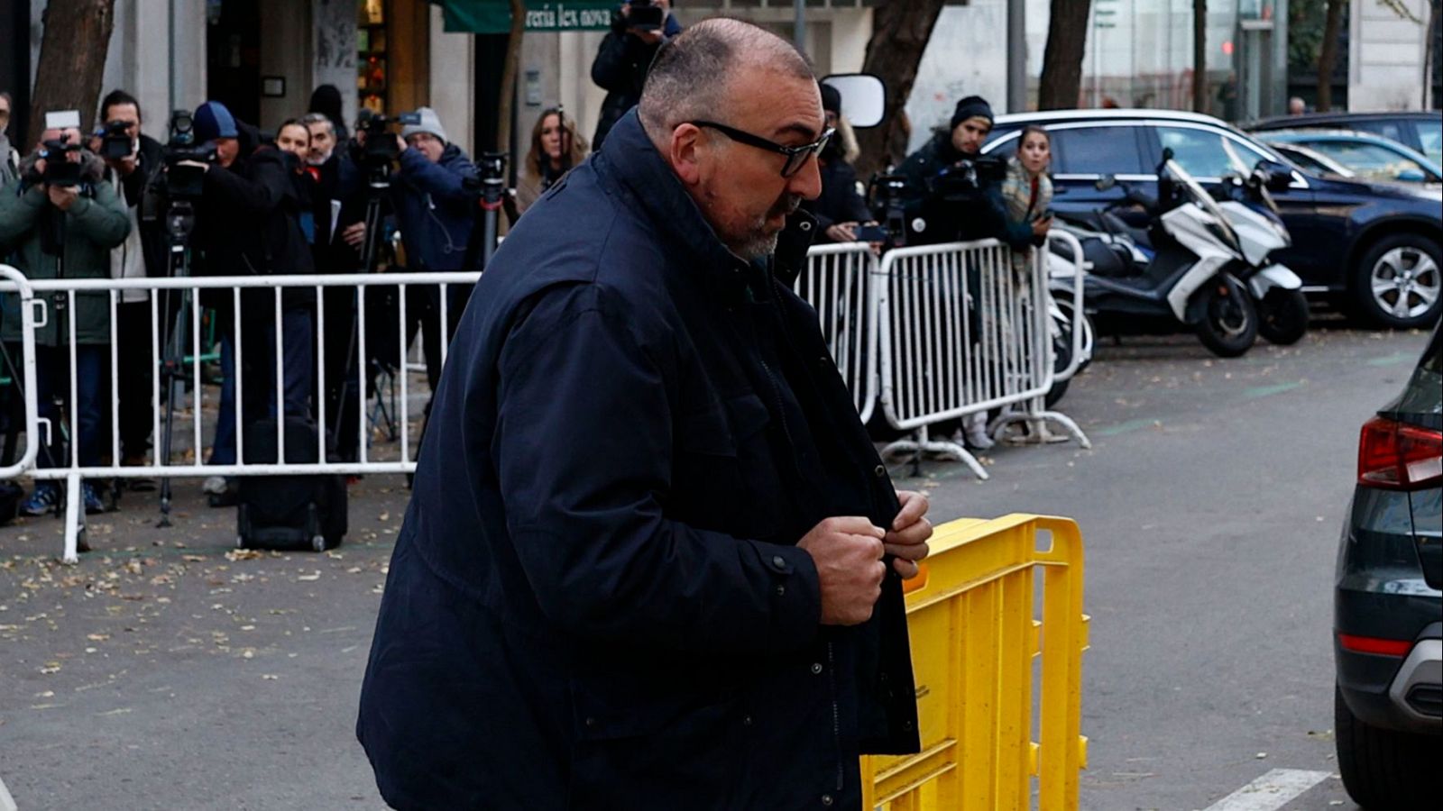 Hombre con chaqueta oscura y gafas sale de una declaración, abriendo una valla amarilla mientras la prensa lo sigue con cámaras y micrófonos.