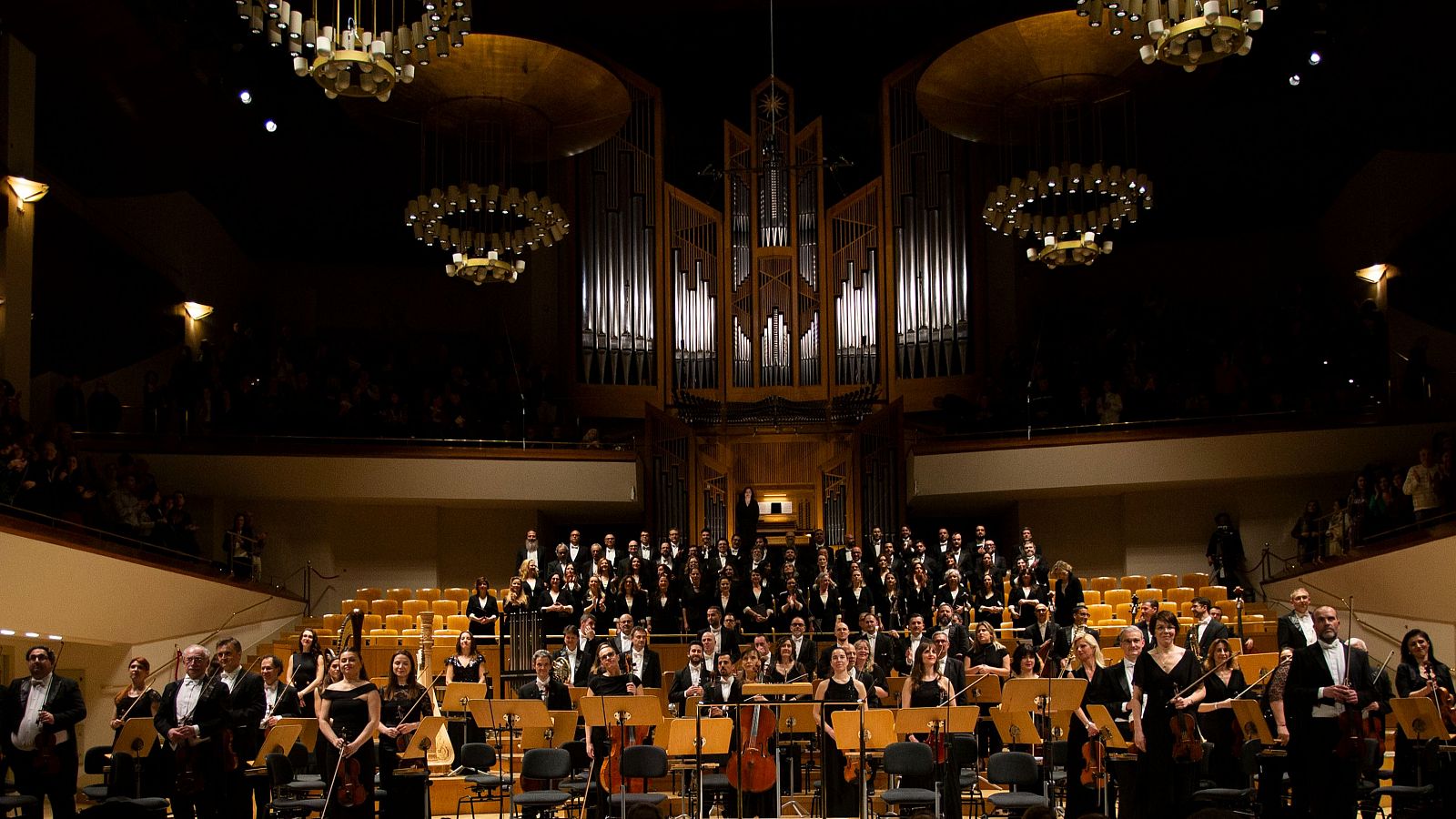 Actuación de la Orquesta Sinfónica y Coro RTVE: vista panorámica del escenario con orquesta, coro y órgano, en un auditorio iluminado. Público presente.