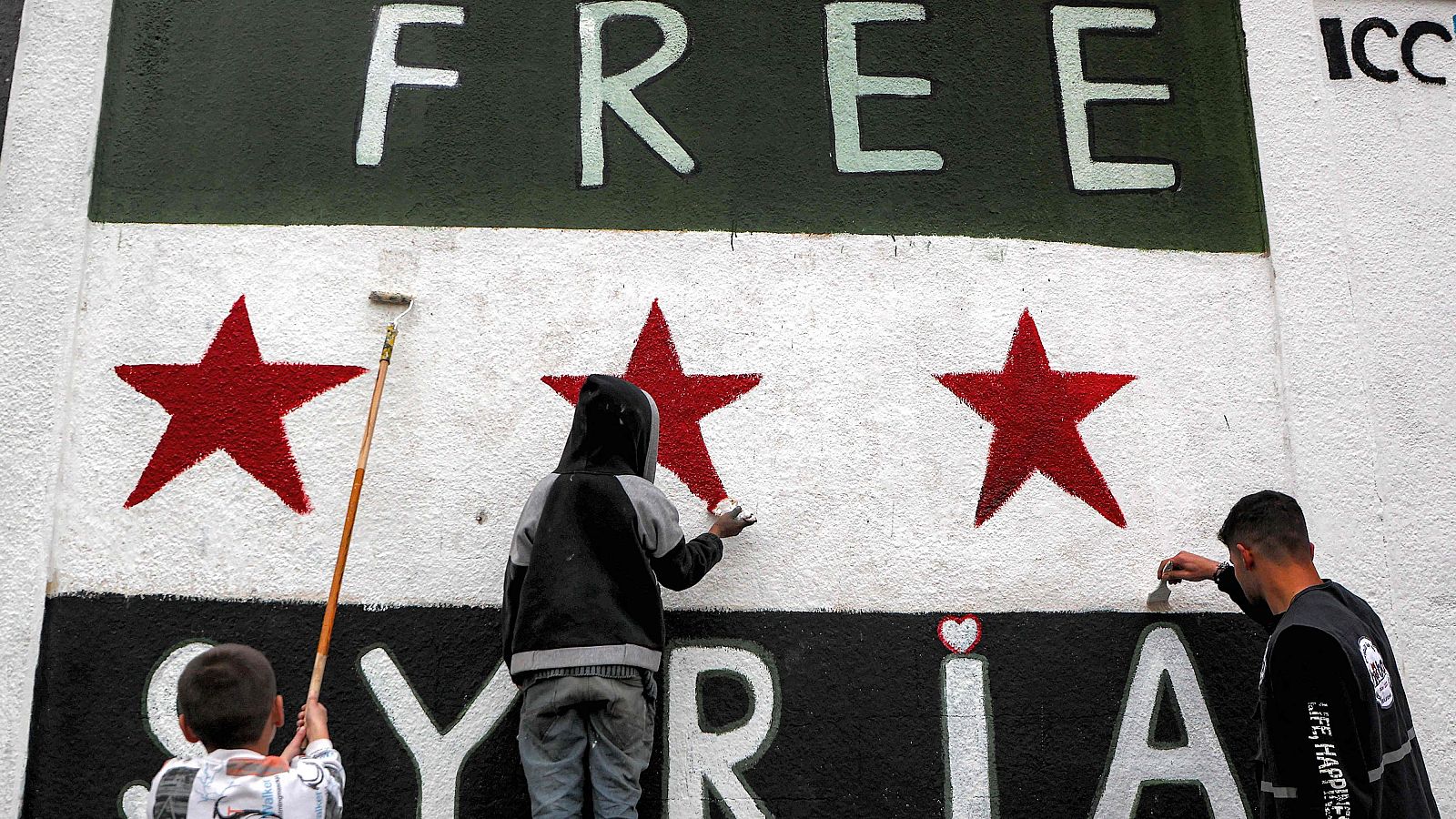 Mural de la bandera siria con las palabras "FREE SYRIA" siendo pintado por tres personas, incluyendo dos niños.  Se observa un mensaje de esperanza y libertad.