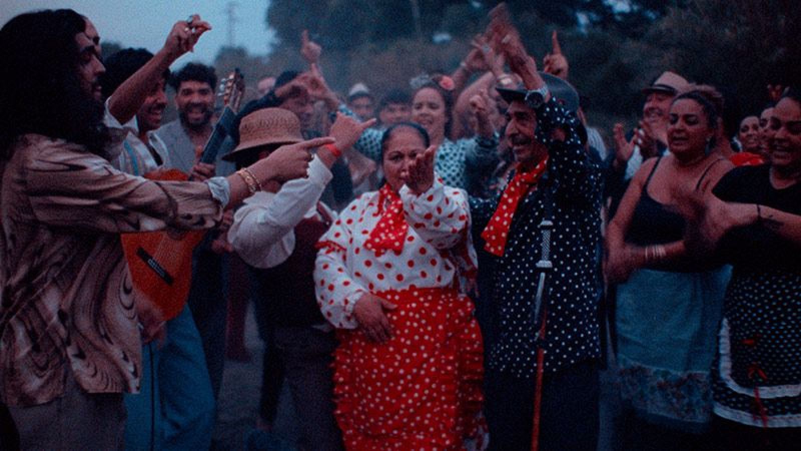 Grupo de personas bailando y tocando música en una celebración nocturna.  Vestidos con lunares, guitarra visible, ambiente festivo.