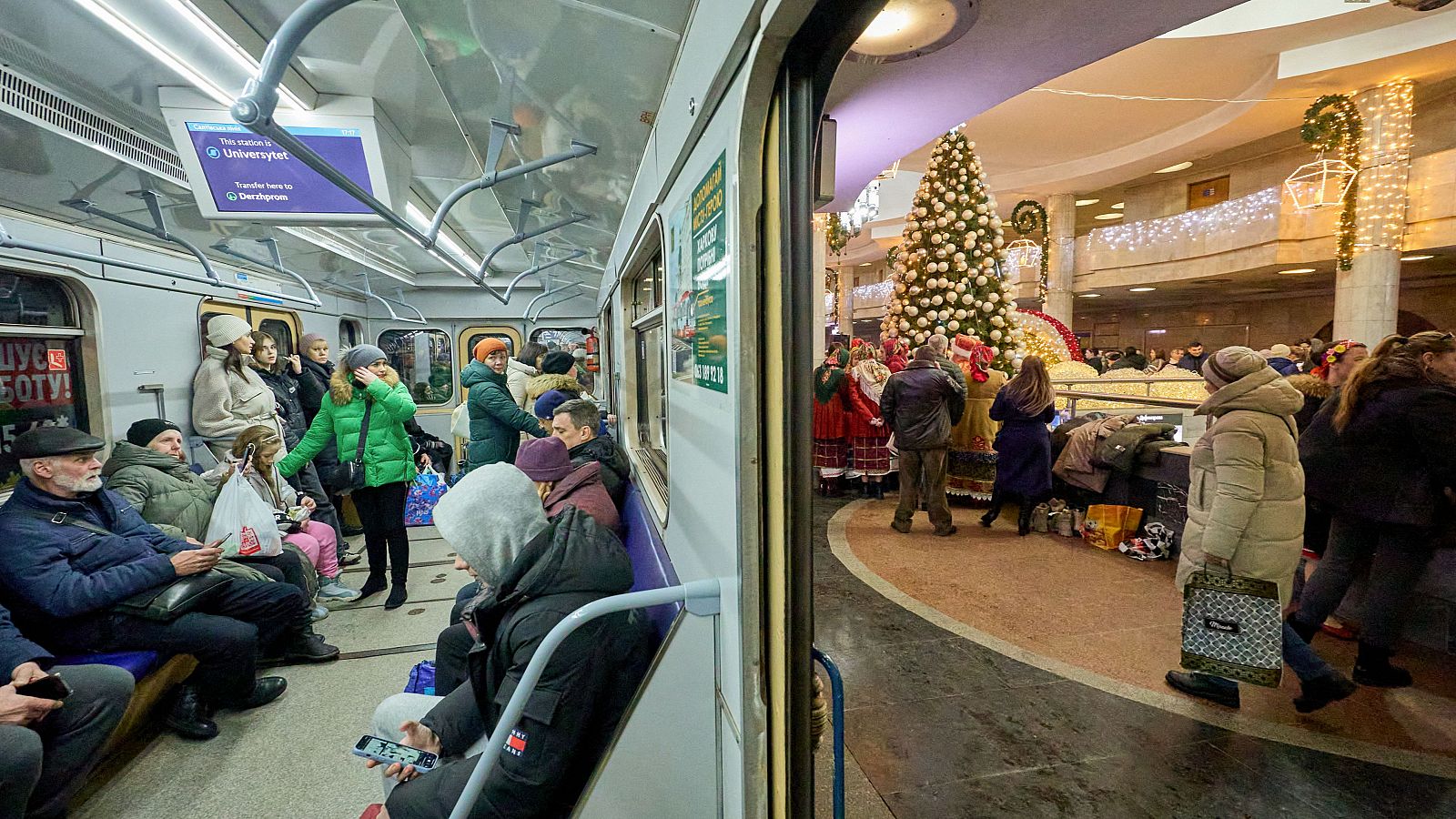 Pasajeros en vagón de metro con cartel digital indicando 'Universytet' y 'Derzhprom'.  Árbol de Navidad decorado en estación, gente reunida alrededor. Escena navideña en contexto bélico.