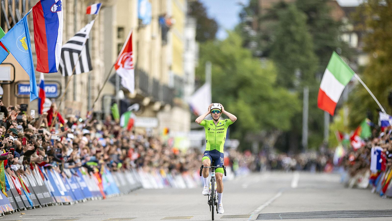 Ciclista con maillot verde lima cruzando la meta, quitándose el casco tras una carrera, rodeado de público que lo celebra.