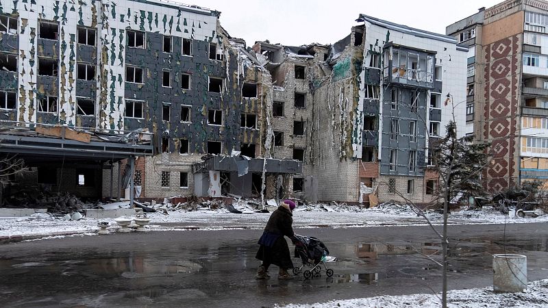 Edificio residencial dañado por bombardeos en invierno.  Una mujer con cochecito pasa cerca, mostrando el impacto de la guerra en la vida civil.