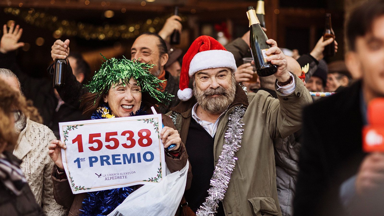 Celebración del primer premio de lotería (15538) con una mujer con tocado verde y un hombre con gorro de Papá Noel. Ambiente festivo navideño.