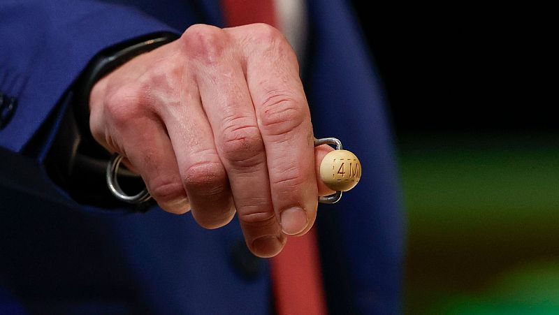 Bola de madera con el número "4M" para el sorteo de Navidad 2024, sostenida por una persona con traje azul oscuro y corbata roja antes de entrar al bombo.