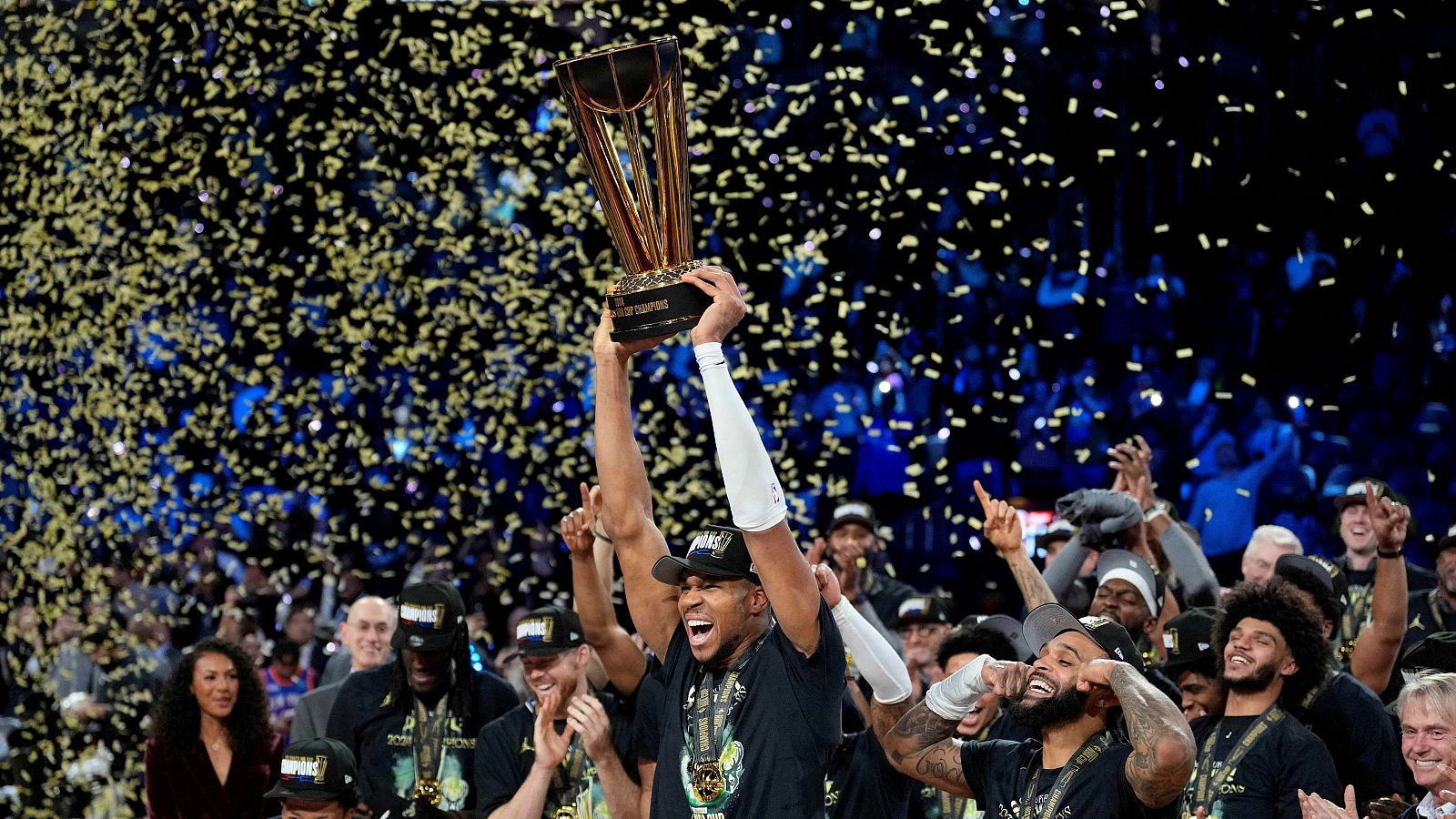 Jugadores de baloncesto celebran victoria con trofeo, confeti dorado y azul, mostrando alegría y euforia.