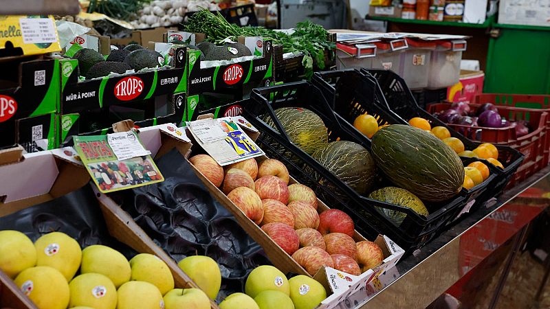 Frutas y verduras frescas en un puesto de mercado, con cajas de manzanas, melones, naranjas y aguacates.  Cajas vacías visibles.