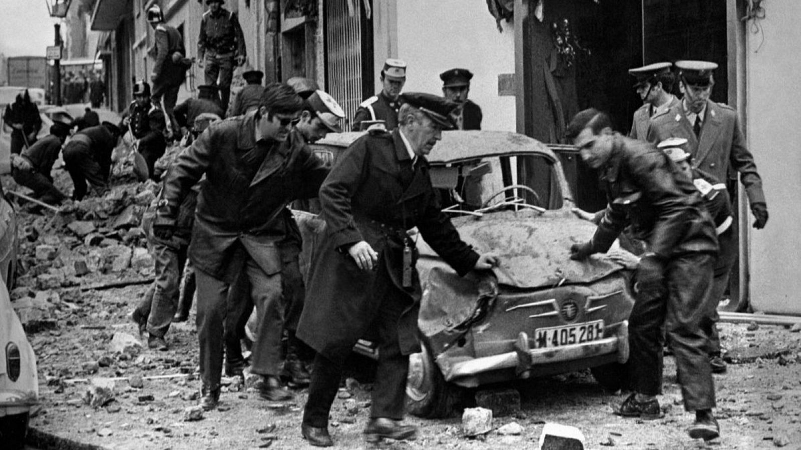 Secuelas de una explosión: coche destrozado (matrícula M-405281), rodeado de personas en uniforme y civiles entre escombros.  Escena caótica en blanco y negro.