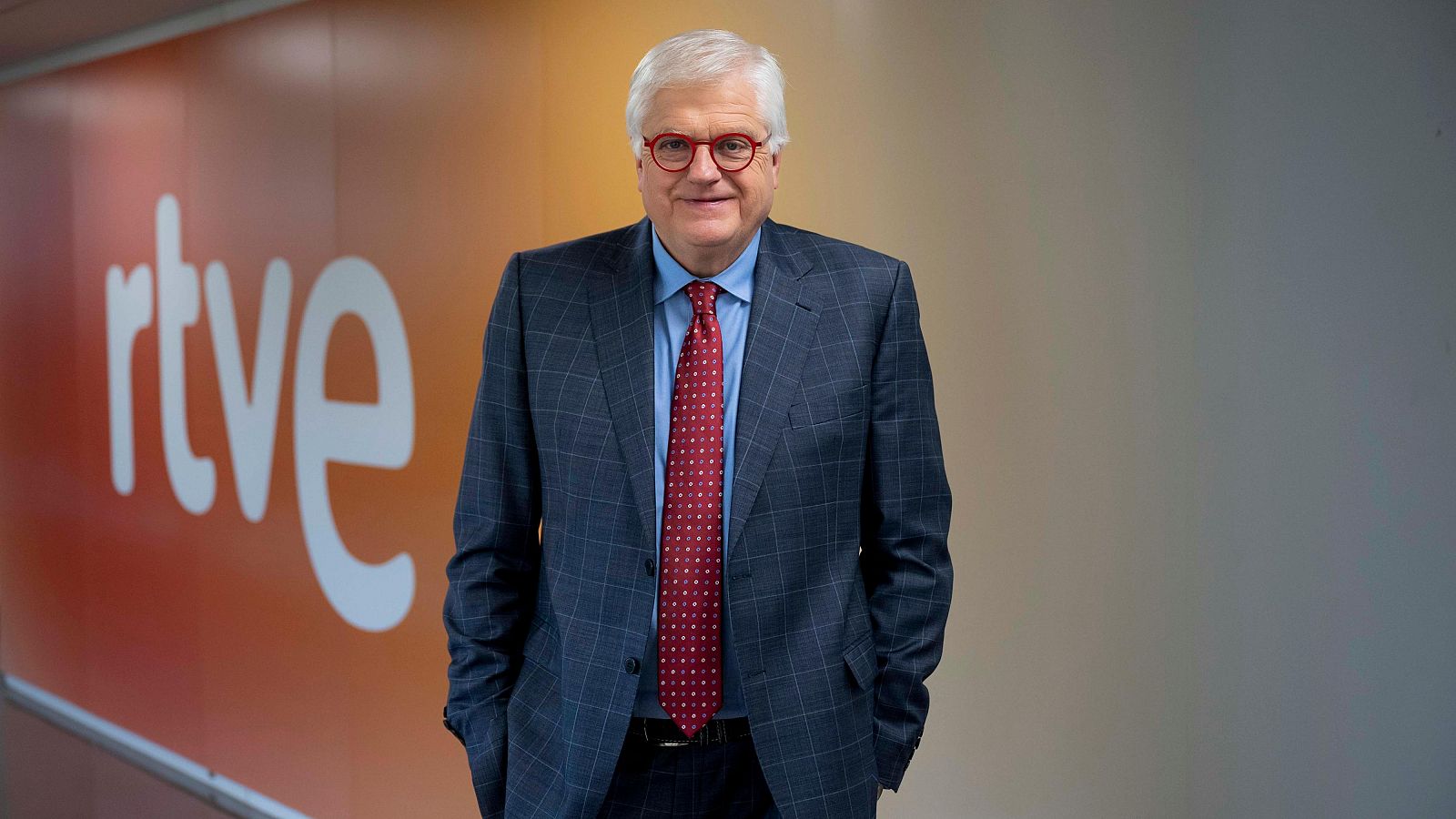 Fotografia de Lluís Falgàs, presentador d''Aquí parlem' de RTVE Catalunya. Vesteix vestit blau fosc a quadres, camisa blau clar i corbata granat.  Es troba dret, amb les mans a les butxaques, davant d'un panell amb el logo de RTVE.