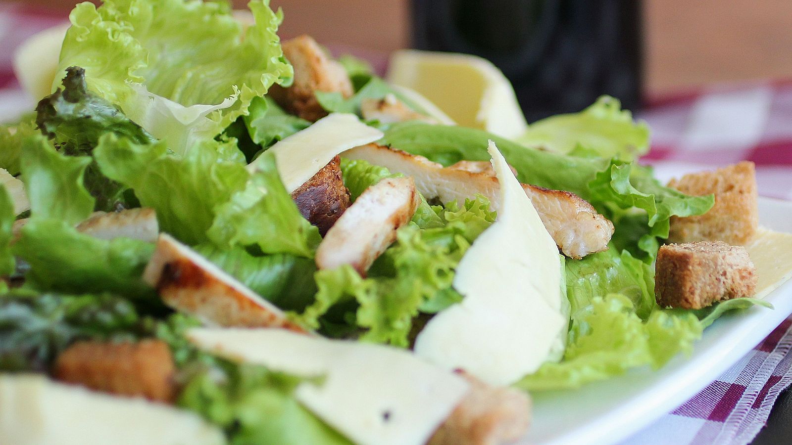 Ensalada César en plato blanco: lechuga, pollo, picatostes y queso parmesano. Fondo con mantel a cuadros y bebida.