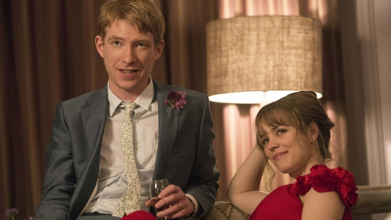 Escena de 'Una cuestión de tiempo': Rachel McAdams en vestido rojo y Domhnall Gleeson en traje gris, sentados en un sofá, sonríen a cámara. Ambiente cálido y festivo.