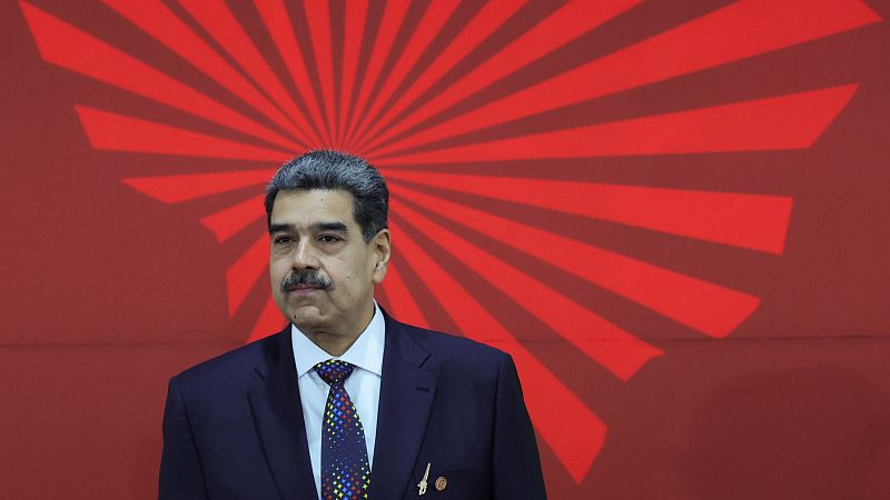 Maduro, en traje oscuro y corbata multicolor, asiste a una cumbre.  Se observa un prendedor en su solapa y un fondo rojo con diseño radial.