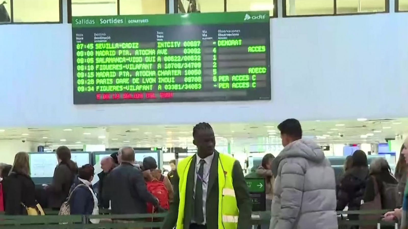 Panel de salidas de trenes con información de hora, destino y vía; se observa retraso en un tren.  Varias personas esperan en la estación, un trabajador asiste a los pasajeros.