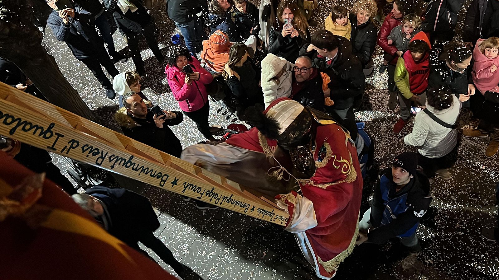 El Patge Faruk sube una escalera de madera en un ambiente festivo, rodeado de gente que le fotografía.  Confeti blanco cubre el suelo.