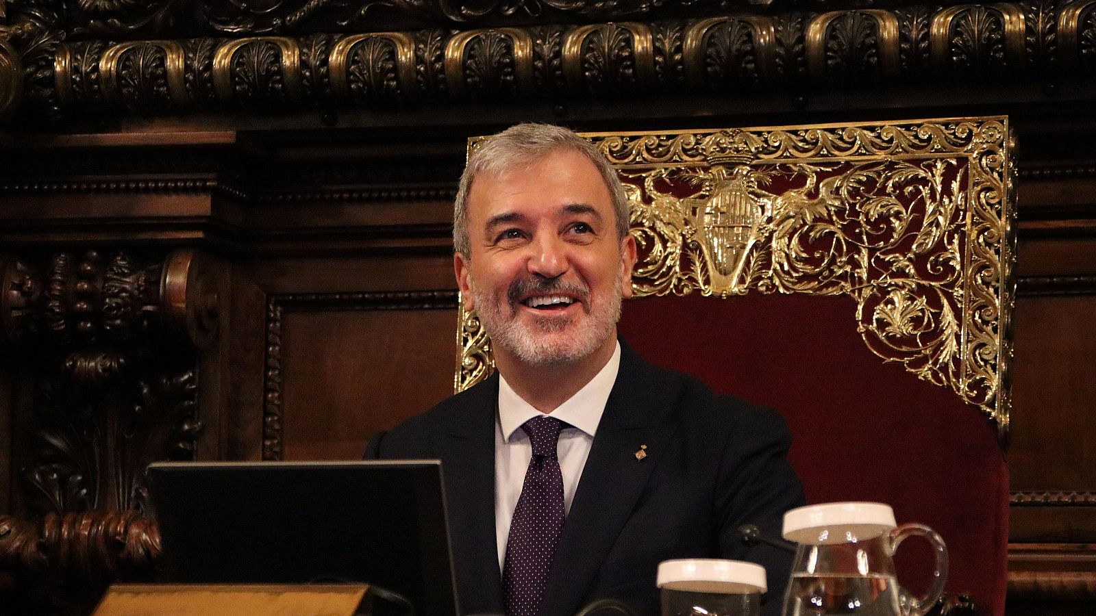 Alcalde en pleno municipal, sentado en silla dorada, traje oscuro y corbata morada. Ambiente formal.