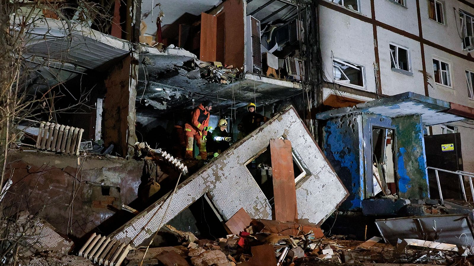 Bomberos inspeccionan los daños de un edificio residencial tras un impacto, con escombros y ventanas rotas. Escena nocturna.