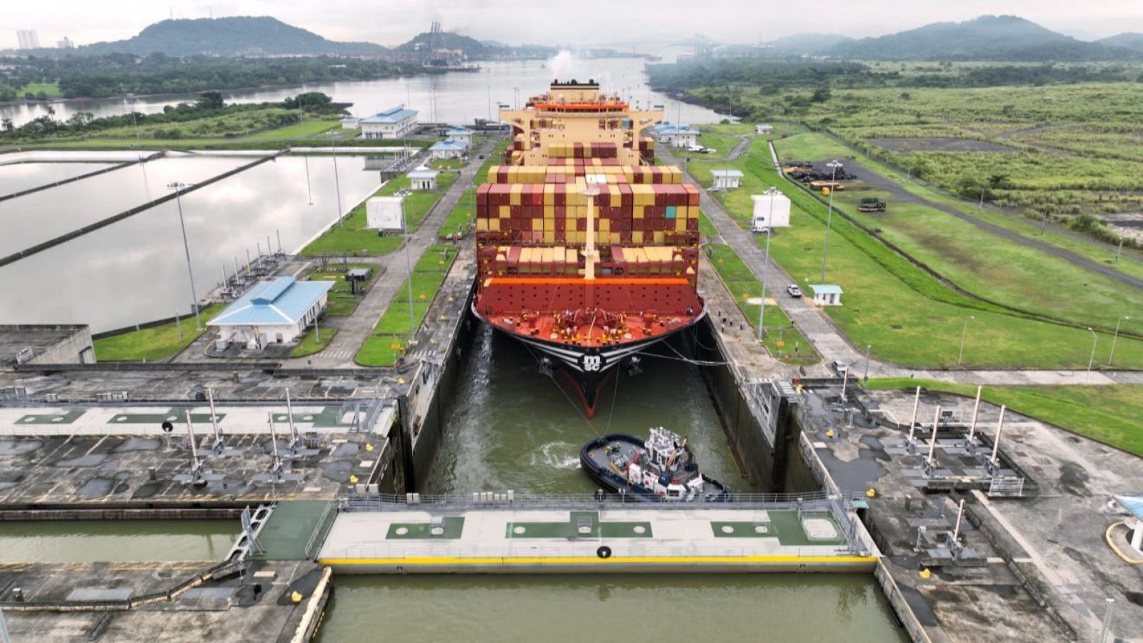 Imagen aérea de un portacontenedores en una esclusa del Canal de Panamá, con un remolcador.  Se aprecian las estructuras de la esclusa y el entorno.