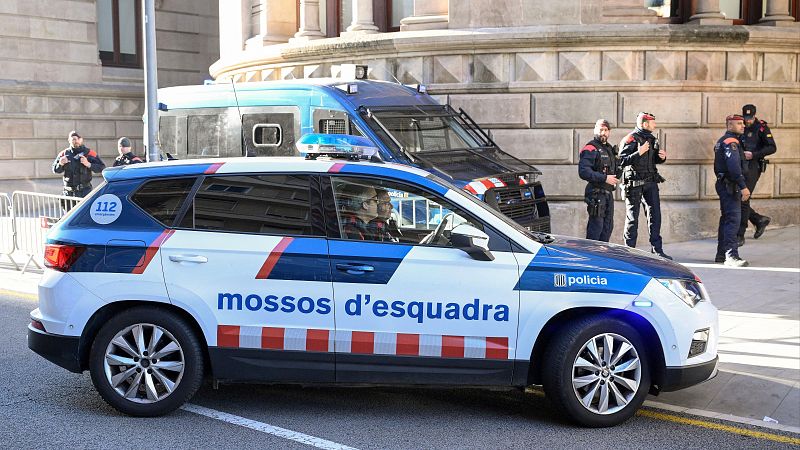 Coche patrulla blanco con franjas rojas y azules aparcado en la calle. Agentes de policía alrededor, algunos con equipo antidisturbios. Imagen de archivo.